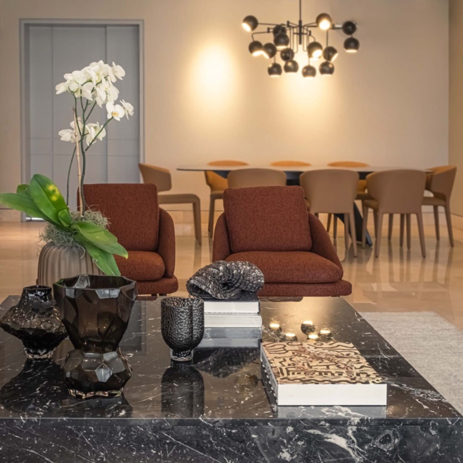 Living room with terracotta chairs and black marble coffee table decorated with flowers and sculptural pieces, dining area in the background with camel chairs and modern pendant lamp
