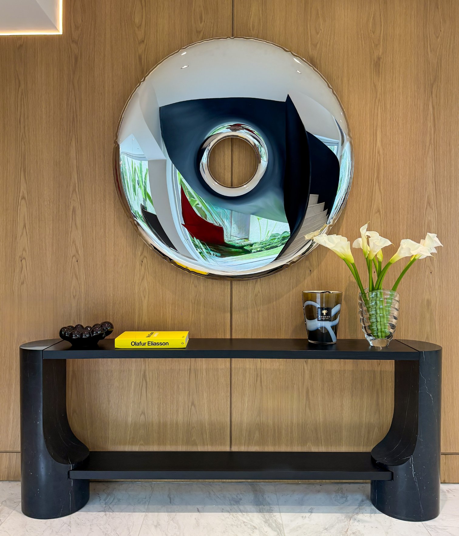 Consola negra con flores blancas y libro de arte Olafur Eliasson bajo espejo circular sobre paneles de madera