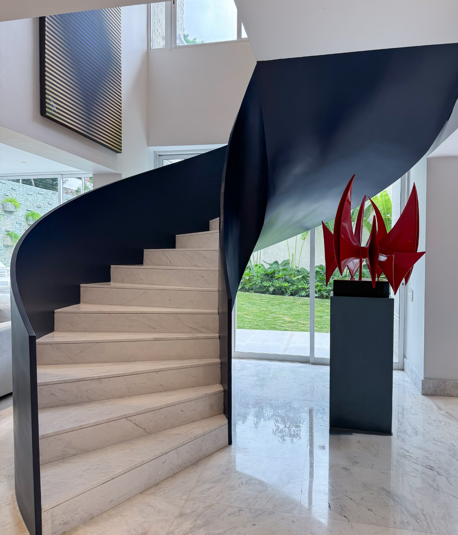 Escalera escultórica con barandilla azul oscuro y escultura roja contemporánea junto a ventanales con vista al jardín