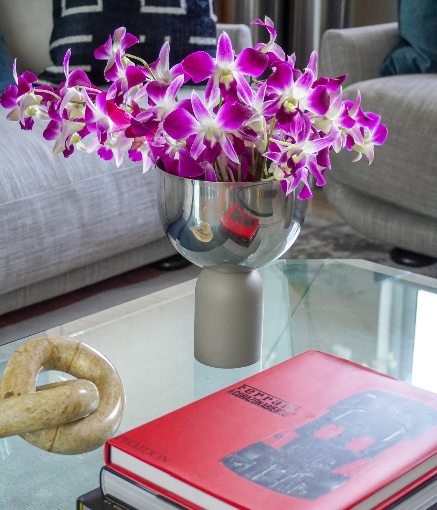 Detalle de mesa de centro de vidrio con libro Ferrari, flores moradas y objetos decorativos